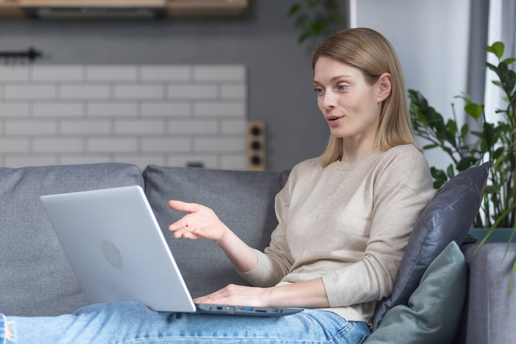Mulher sentada em casa no sofá da cozinha, usando um laptop para uma chamada de vídeo, conversando com amigos remotamente - Foto, Imagem