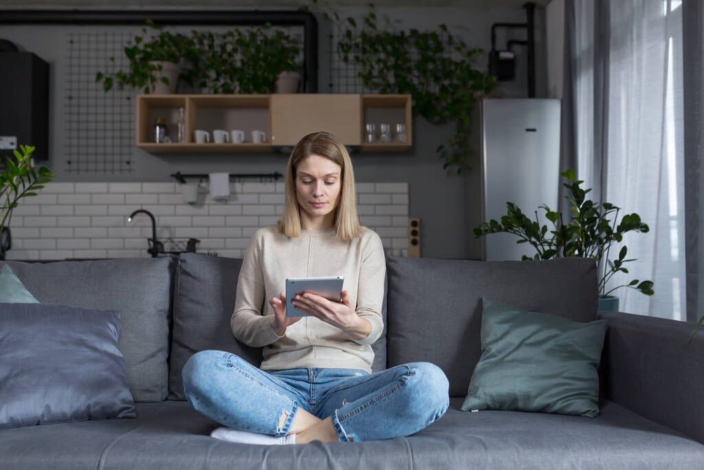 Mujer sentada en un sofá en casa, usando tableta, leyendo libros electrónicos y aprendiendo en línea - Foto, Imagen