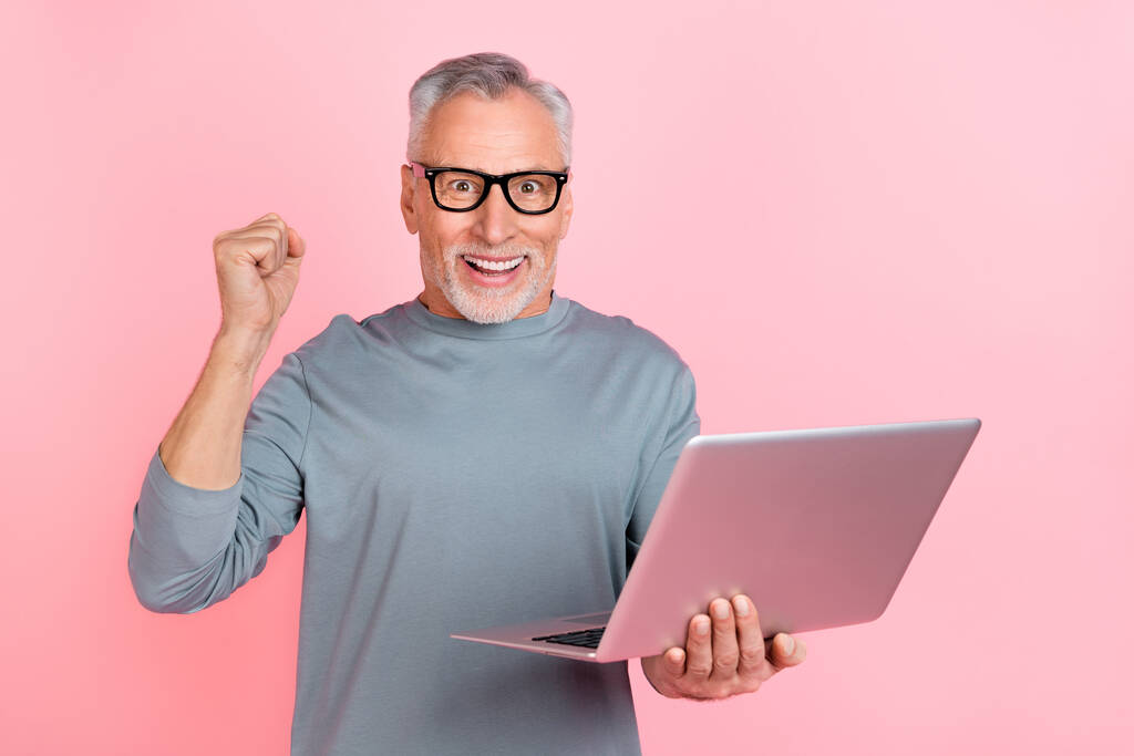 Foto von cleveren Glückspilz Rentner gekleidet graues Hemd Brille Lesen modernes Gerät steigt Faust isoliert rosa Hintergrund - Foto, Bild