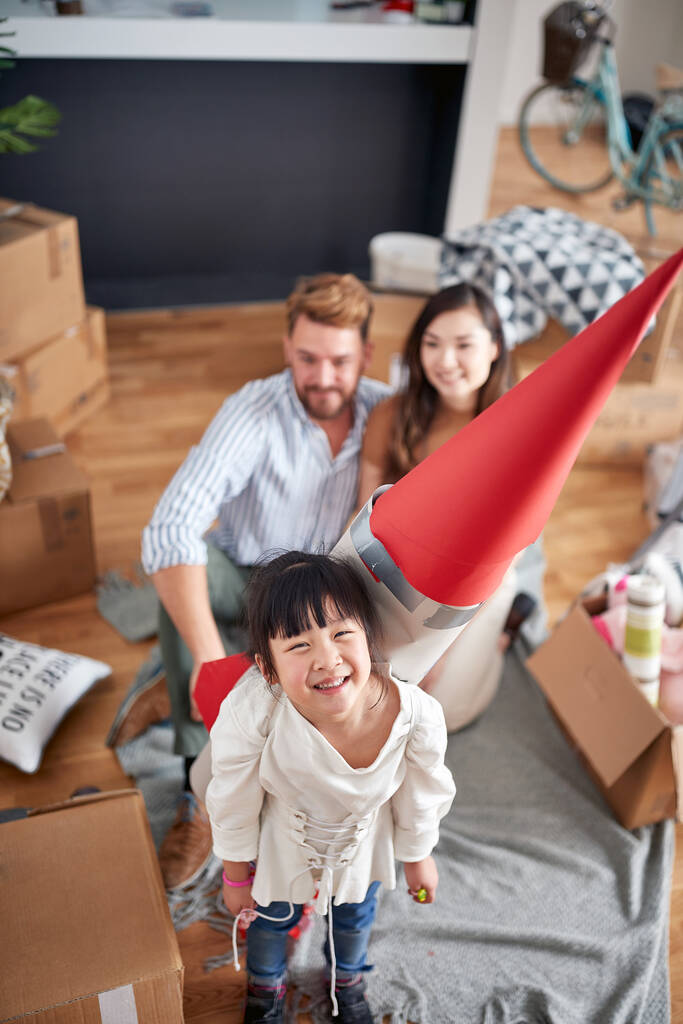 Вид сверху на счастливую маленькую девочку, играющую со своими родителями с бумажной ракетой на спине. многонациональная семья в новой квартире - Фото, изображение