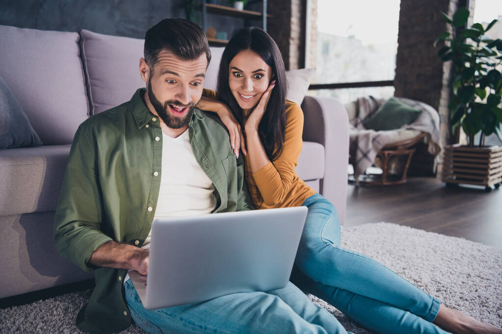 Portrait de beau beau couple joyeux étonné assis sur le tapis en utilisant un ordinateur portable à la maison appartement à l'intérieur - Photo, image