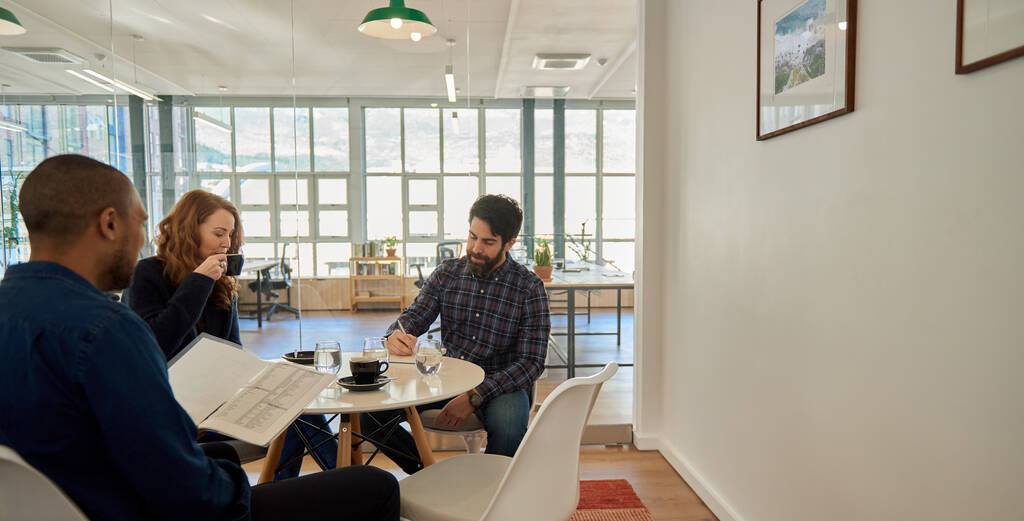 Tre giovani imprenditori che parlano di un progetto insieme durante un incontro davanti a un caffè intorno a un tavolo in un ufficio - Foto, immagini