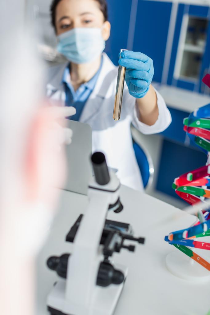 genetista offuscata in maschera medica che tiene la provetta vicino al microscopio e al modello di DNA - Foto, immagini