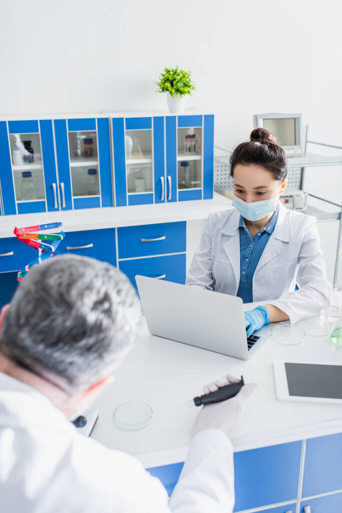 scientist in medical mask working on laptop near digital tablet with blank screen and blurred colleague - Photo, Image