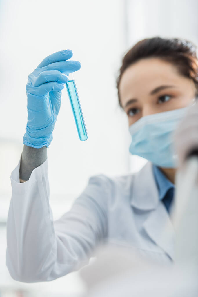 Tıbbi maskeli bulanık bir kadın mavi sıvıyla test tüpüne bakıyor. - Fotoğraf, Görsel
