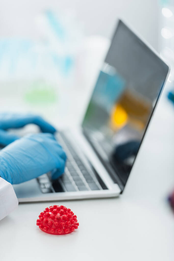 μερική άποψη του ιολόγου δακτυλογράφηση σε φορητό υπολογιστή με κενή οθόνη κοντά στο κόκκινο coronavirus βακτηριακό μοντέλο - Φωτογραφία, εικόνα