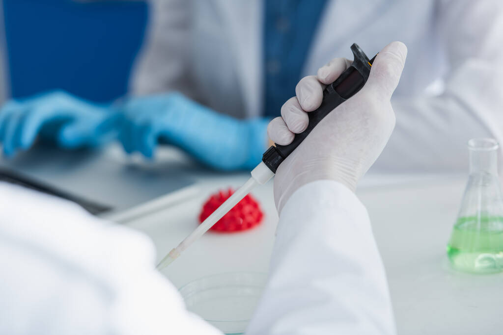 oříznutý pohled vědce v latexové rukavici držící mikropipetku v blízkosti rozmazaného modelu koronavirových bakterií - Fotografie, Obrázek