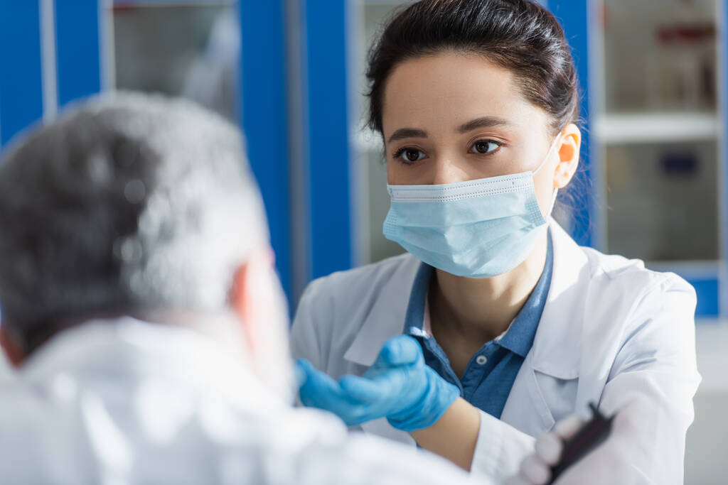 биоинженер в медицинской маске и латексной перчатке указывает рукой, разговаривая с размытым коллегой - Фото, изображение
