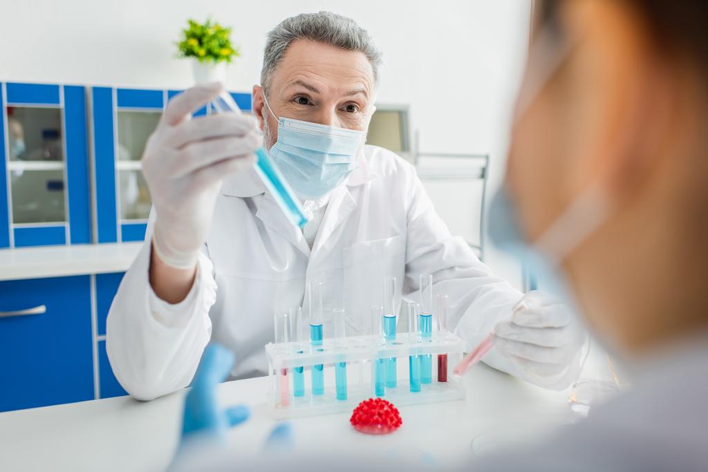 Bioingenieur in medizinischer Maske im Reagenzglas neben verschwommenem Kollegen und Coronavirus-Bakterienmodell - Foto, Bild