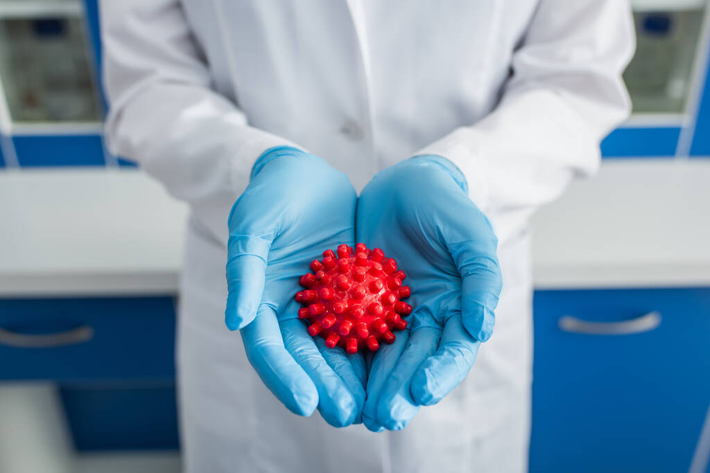vista recortada del biólogo en guantes de látex azul que sostiene el modelo de bacteria del coronavirus rojo  - Foto, Imagen