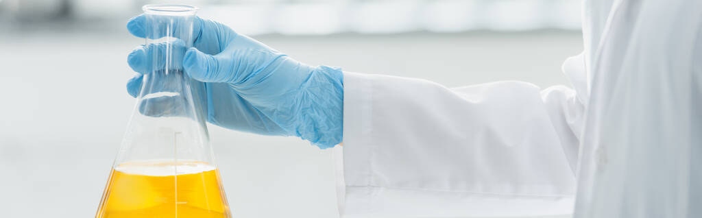 Teilansicht des Wissenschaftlers im Latexhandschuh mit medizinischem Kolben mit gelber Flüssigkeit, Banner - Foto, Bild
