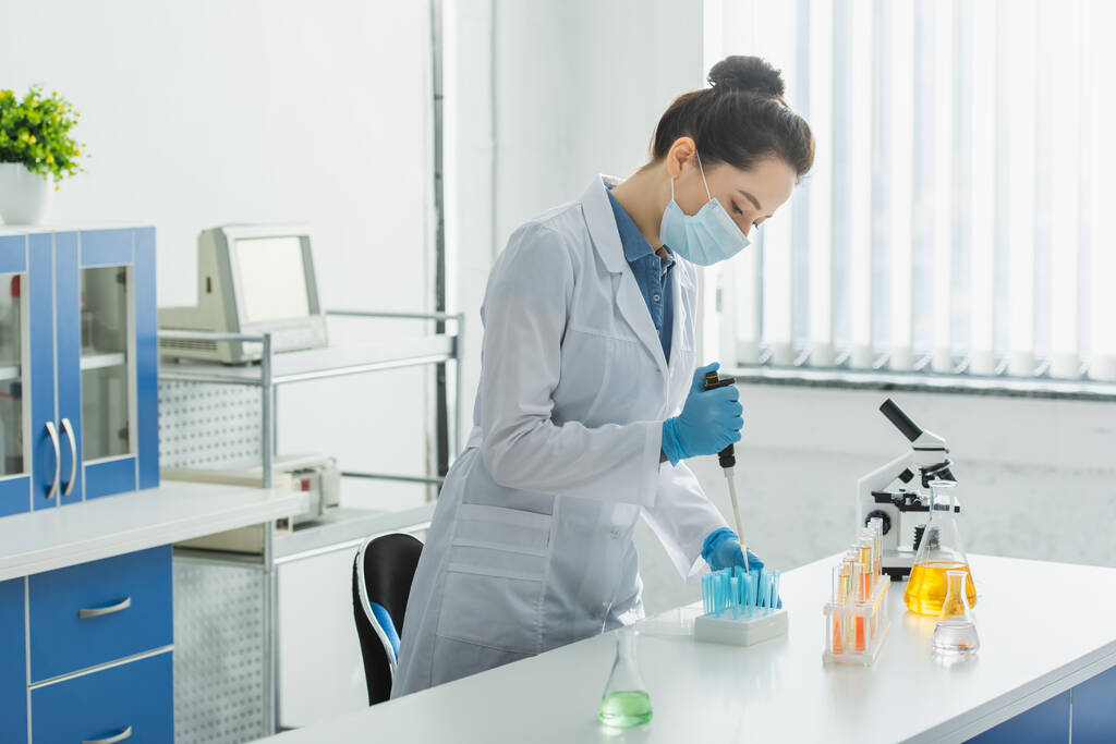 mikropipetillä ja koeputkilla mikroskoopin lähellä työskentelevä lääketieteellisen naamion ja valkoisen takin bioinsinööri - Valokuva, kuva