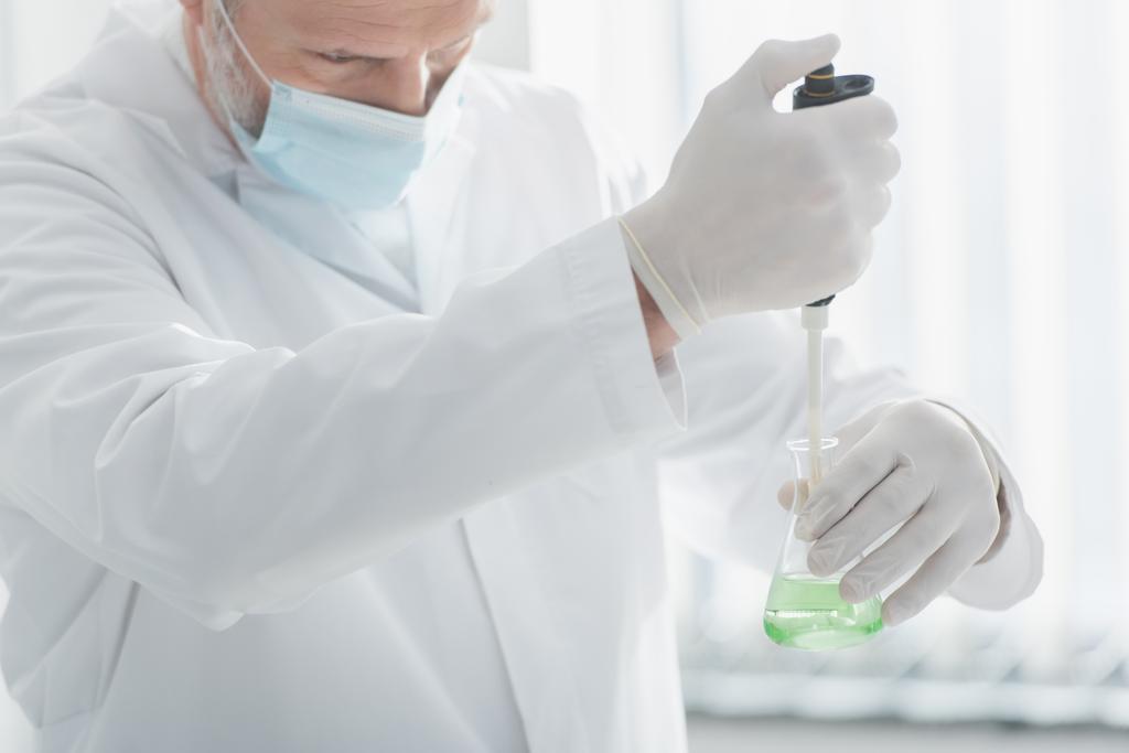 вчений в медичній масці і латексних рукавичках тримає мікроплівку і колбу з рідиною
 - Фото, зображення