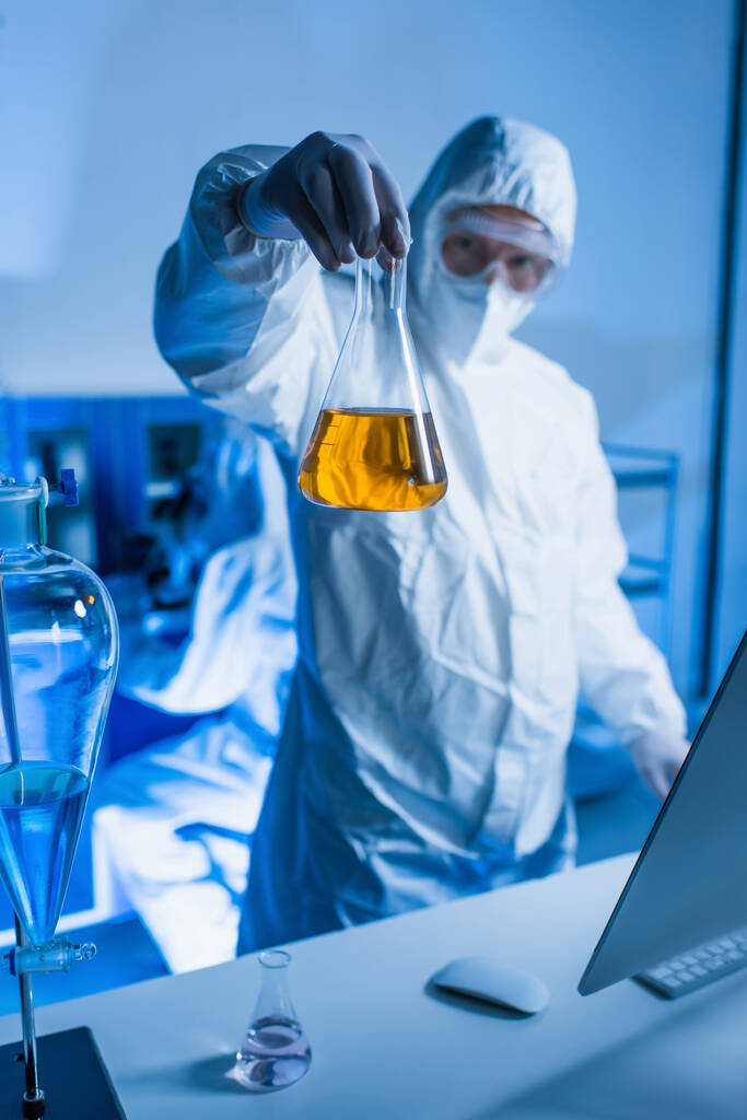 研究室でオレンジの液体でフラスコを保持するハズマットスーツのぼやけた科学者 - 写真・画像