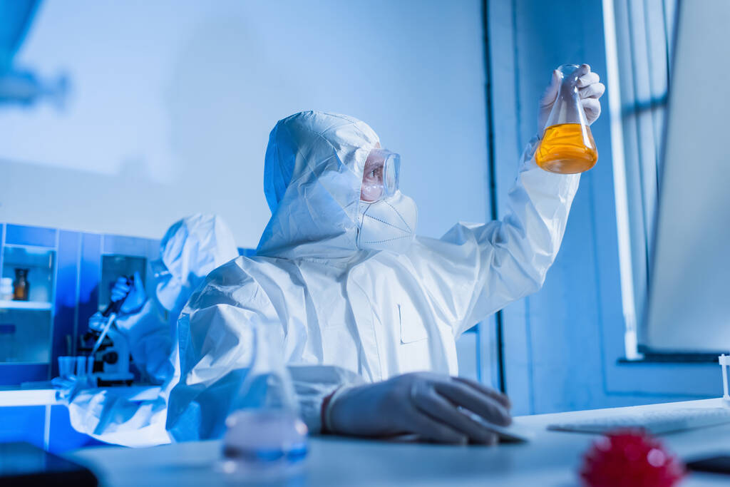bio-ingenieur in hazmatpak op zoek naar kolf met sinaasappelvloeistof in kolf - Foto, afbeelding