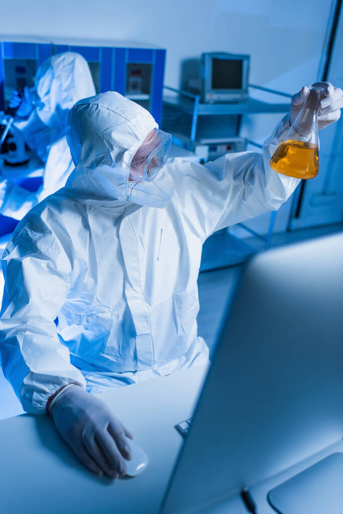 vědec v ochranném obleku drží baňku s oranžovou kapalinou v blízkosti rozmazaného monitoru v laboratoři - Fotografie, Obrázek