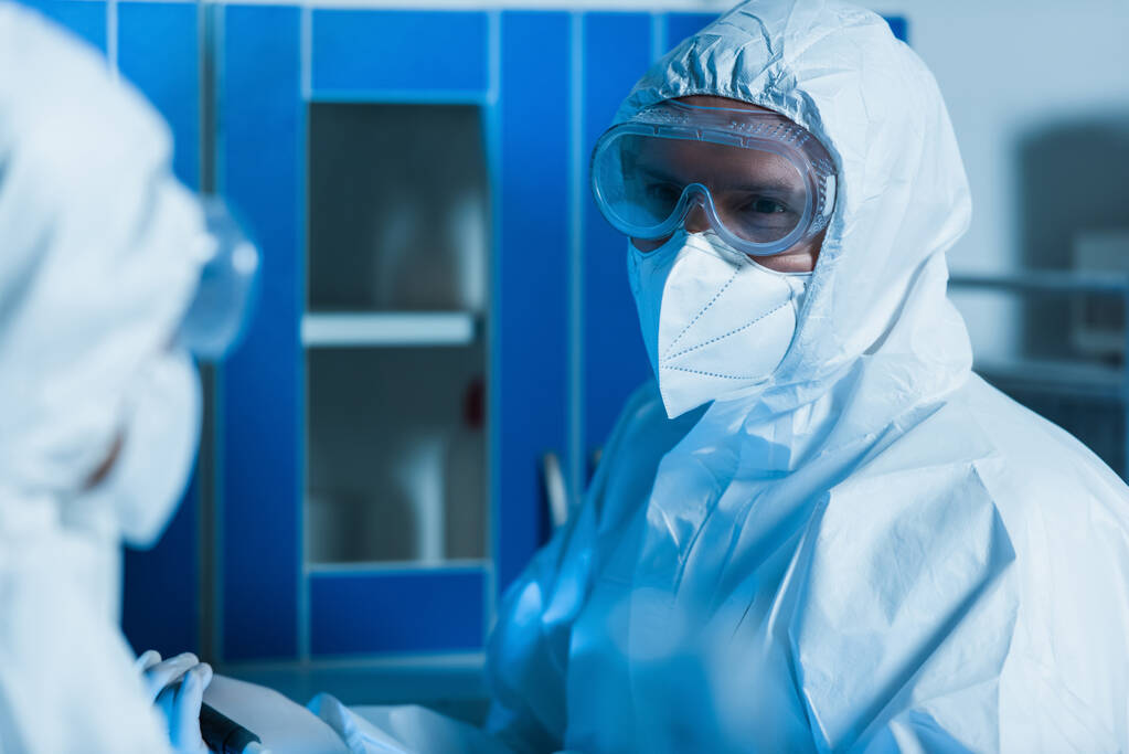 βιομηχανικός με στολή, γυαλιά και ιατρική μάσκα που κοιτάζει συνάδελφο σε θολή πρώτο πλάνο - Φωτογραφία, εικόνα