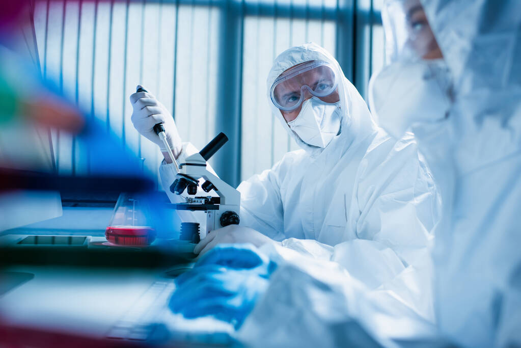 mężczyzna w goglach i masce medycznej w pobliżu mikroskopu i naukowiec na zamazanym pierwszym planie - Zdjęcie, obraz