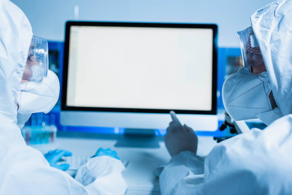 vědec v ochranném obleku a lékařské masce ukazuje na monitor s bílou obrazovkou při práci s kolegou - Fotografie, Obrázek