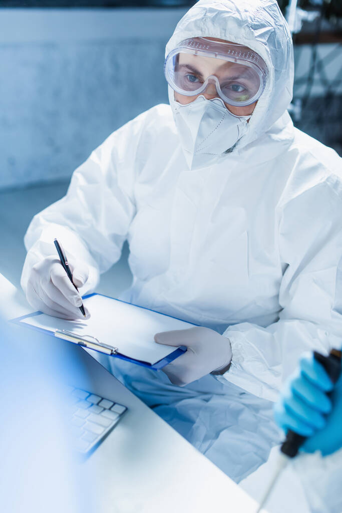 Tehlikeli madde giysisi içindeki genetikçi elinde boş bir not tahtası ve kalemle laboratuvarda bekliyor. - Fotoğraf, Görsel