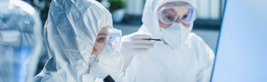 биоинженер в персональном защитном оборудовании рядом с коллегой указывая на размытый фон, баннер - Фото, изображение