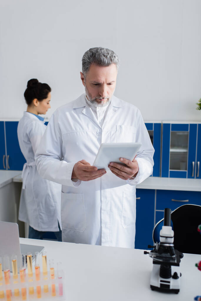 naukowiec w średnim wieku patrząc na cyfrową tabletkę podczas pracy w pobliżu kolegi w laboratorium - Zdjęcie, obraz