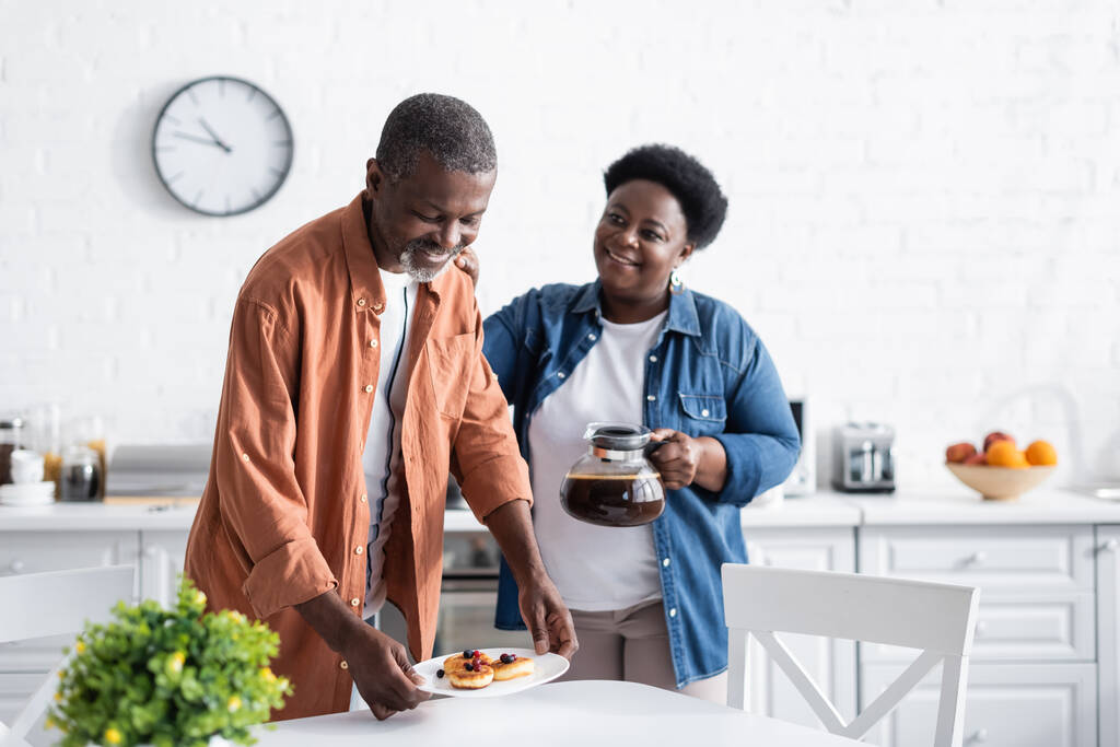 glücklicher afrikanisch-amerikanischer Mann hält Pfannkuchen auf Teller neben Frau mit Kaffeekanne - Foto, Bild