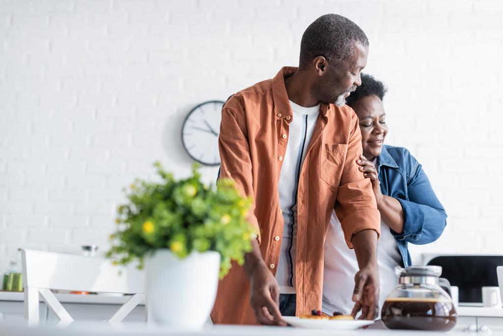 glücklich senior afrikanisch amerikanisch frau umarmt ehemann in der nähe frühstück in küche  - Foto, Bild