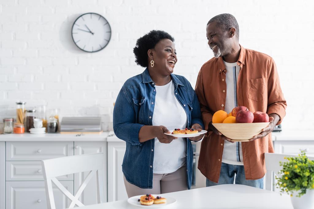 счастливый старший африканский американец держит миску с фруктами рядом с улыбающейся женой - Фото, изображение