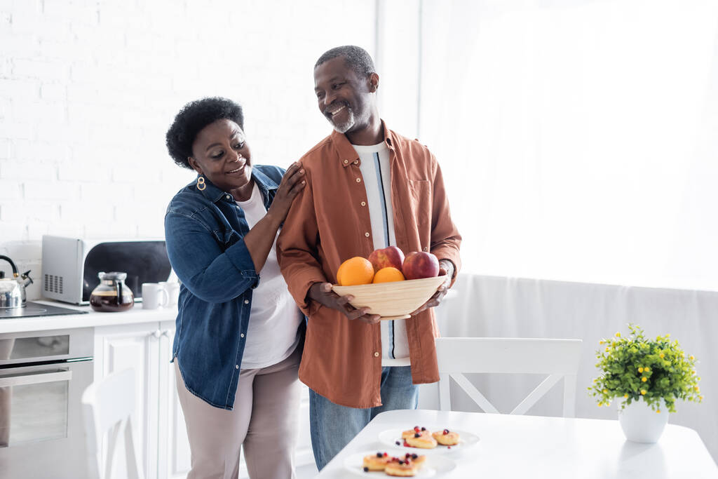 sénior afroamericano hombre celebración bowl con frutas frescas cerca sonriente esposa en cocina  - Foto, imagen