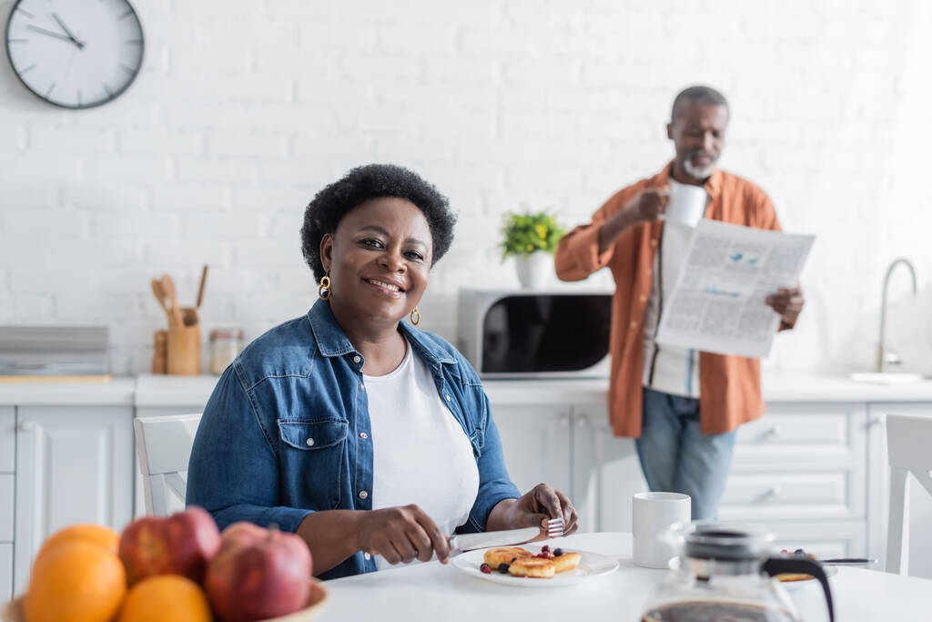 boldog és idős afro-amerikai nő reggelizik közelében elmosódott férj újsággal - Fotó, kép