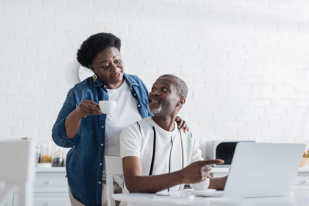 Sakallı Afrikalı Amerikalı adam dizüstü bilgisayarı işaret edip karısına bakıyor. - Fotoğraf, Görsel