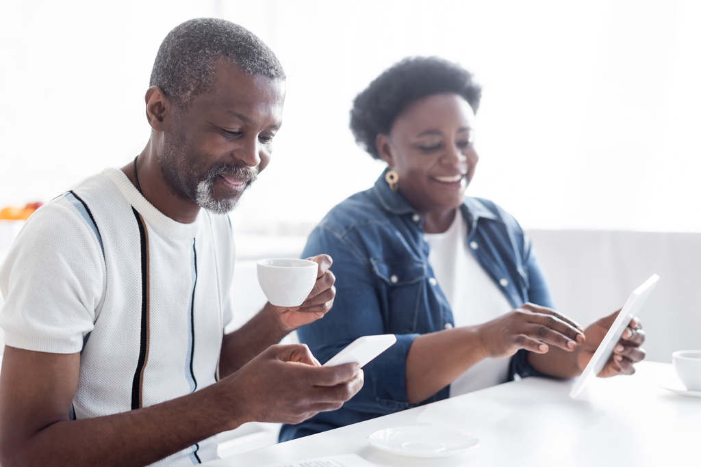 senior africký Američan muž s šálkem kávy pomocí smartphone v blízkosti rozmazané manželky s digitálním tabletem - Fotografie, Obrázek