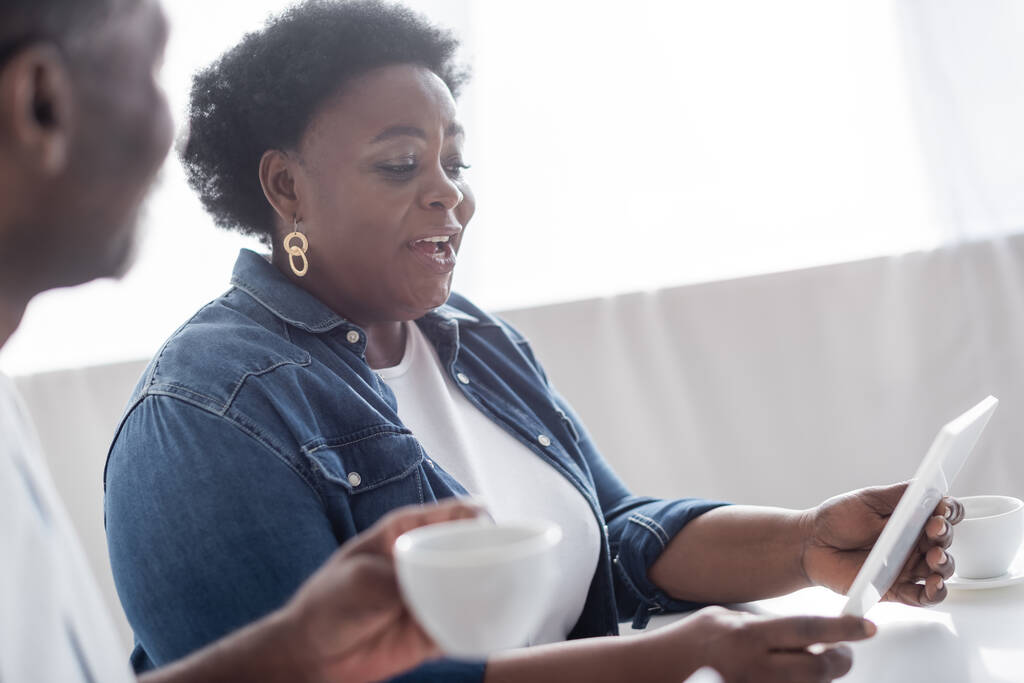 Senior afrikanisch-amerikanische Frau hält digitales Tablet neben verschwommenem Ehemann mit Tasse Kaffee - Foto, Bild