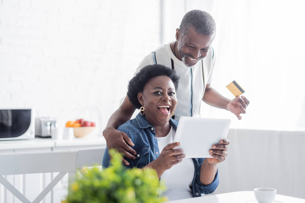ανώτερος αφρικανός Αμερικανός άνδρας κρατώντας πιστωτική κάρτα κοντά κατάπληκτος σύζυγος με ψηφιακή ταμπλέτα  - Φωτογραφία, εικόνα
