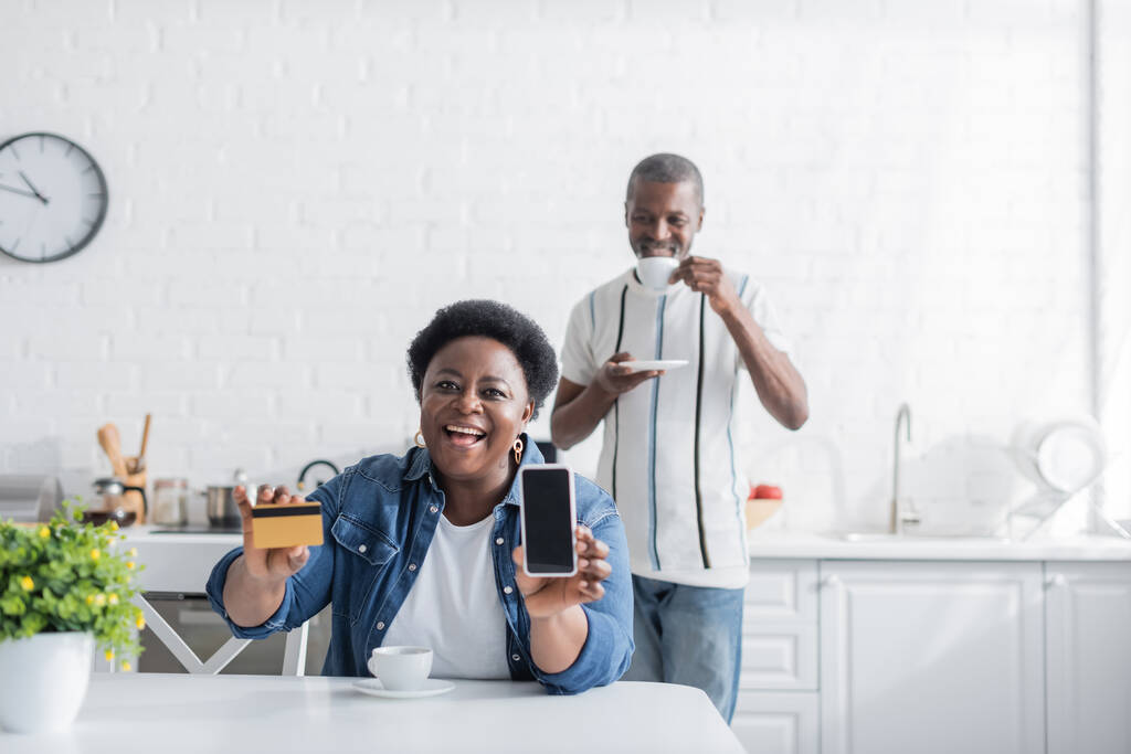 feliz adulto mayor afroamericana mujer sosteniendo tarjeta de crédito y teléfono inteligente con pantalla en blanco cerca del marido  - Foto, imagen