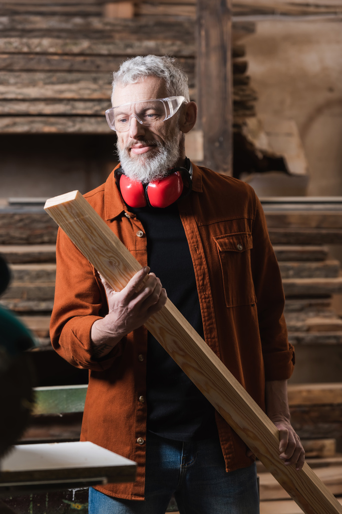 Gözlüklü olgun mobilya tasarımcısı marangozluk stüdyosunda ahşap kalas tutuyor. - Fotoğraf, Görsel