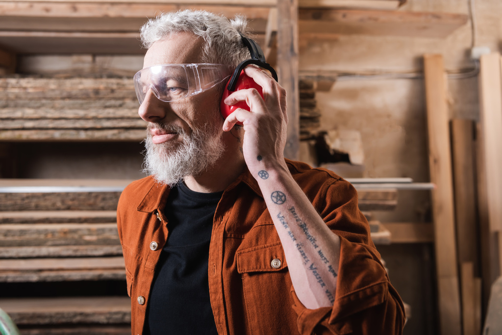 Gözlüklü dövmeli marangoz atölyedeki koruyucu kulaklıkları ayarlıyor. - Fotoğraf, Görsel
