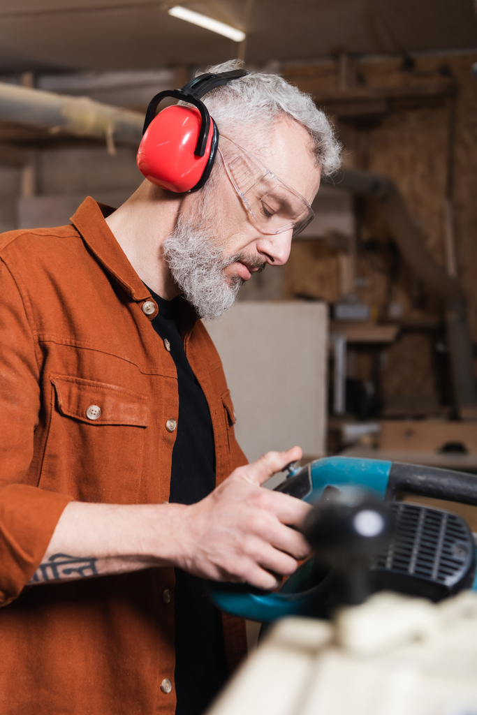 Tischler mit schützenden Ohrenschützern arbeitet in Werkstatt mit Gehrungssäge - Foto, Bild