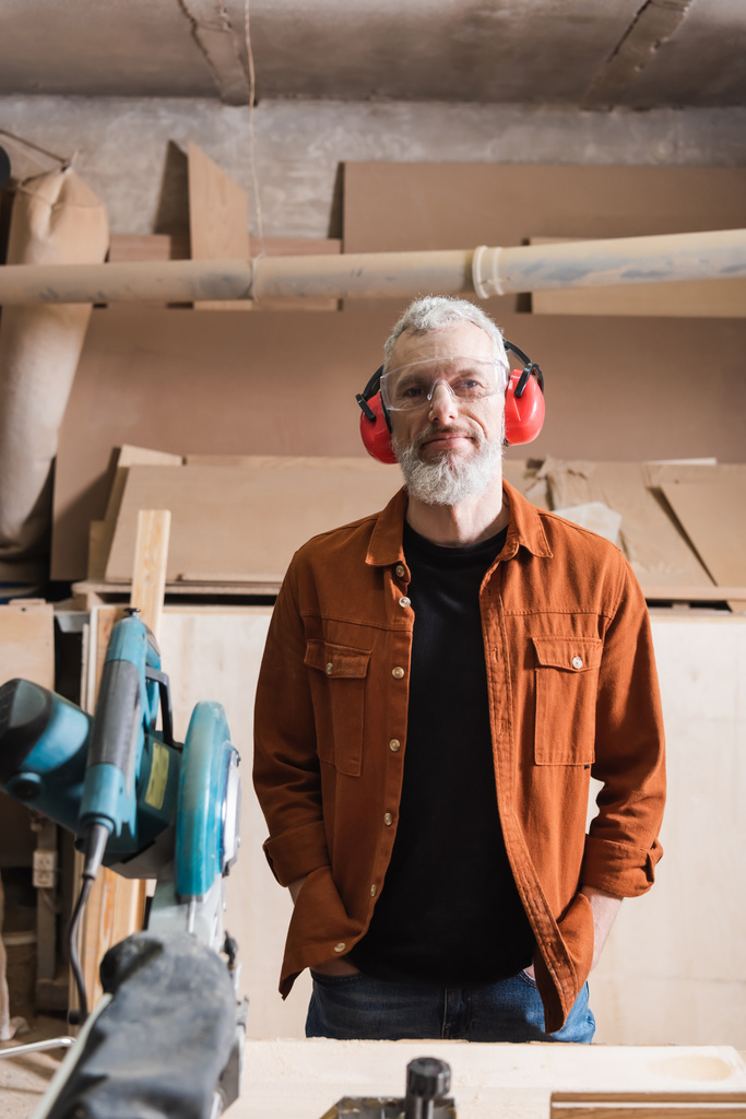 volwassen meubelontwerper in bril en oorkappen staand met handen in zakken in werkplaats - Foto, afbeelding
