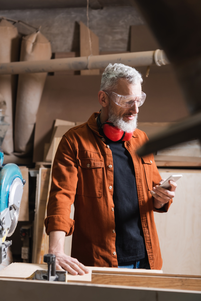 bearded furniture designer in goggles using smartphone on blurred foreground - Photo, Image