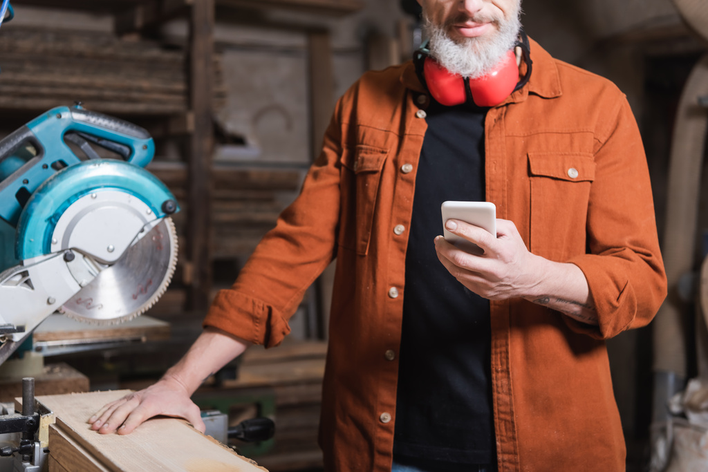обрізаний вид бородатого дерев'яного працівника з мобільним телефоном поруч з мітмером побачив у майстерні
 - Фото, зображення