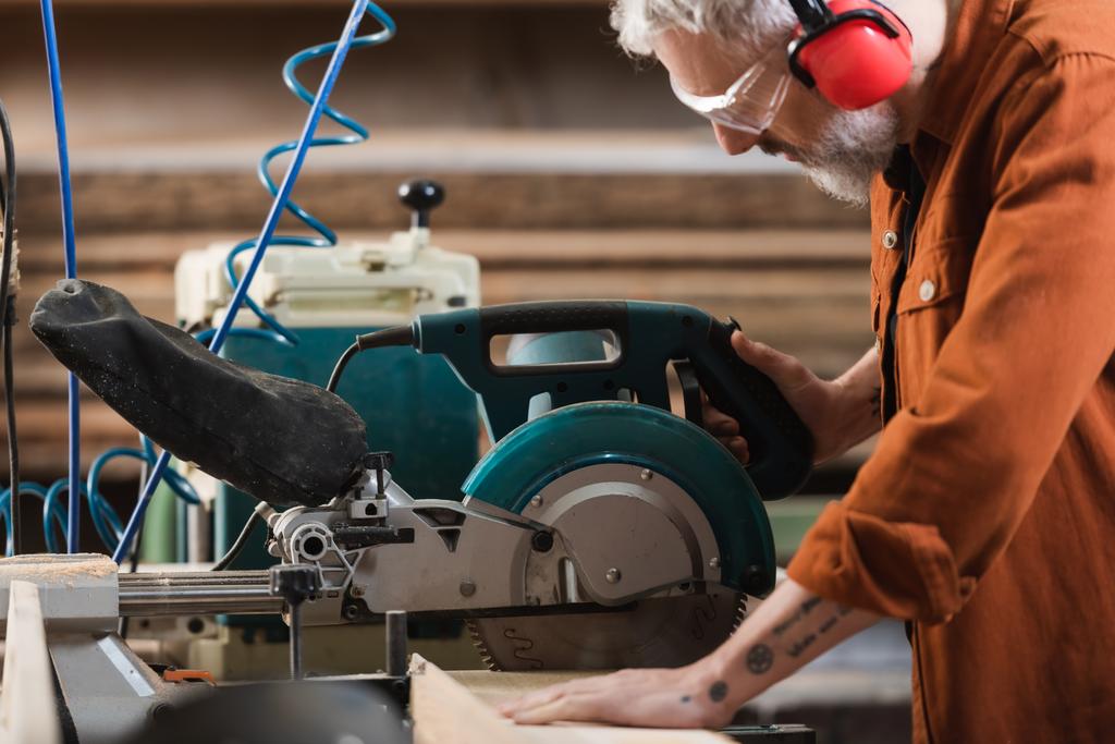 vista lateral de carpintero de mediana edad cortando madera en sierra de inglete - Foto, imagen
