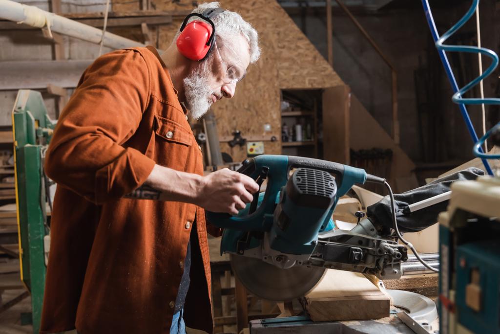 šedovlasý tesař pracující s mitrovou pilou v dřevoobráběcím studiu - Fotografie, Obrázek