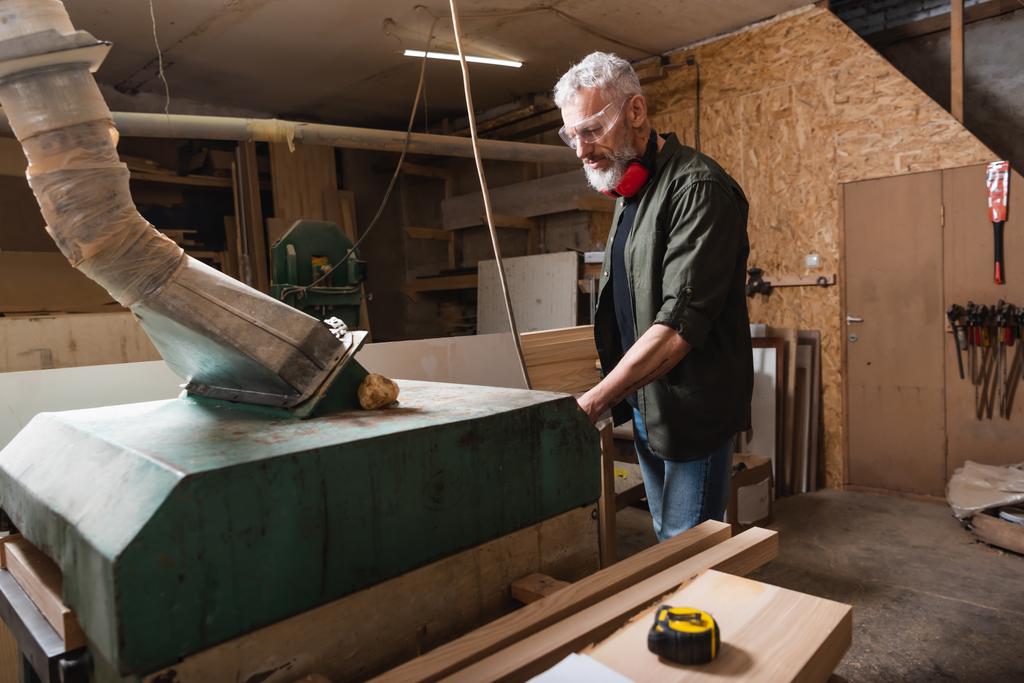grijsharige houtbewerker werkt in de buurt van ventilatie kap in timmerwerk studio - Foto, afbeelding
