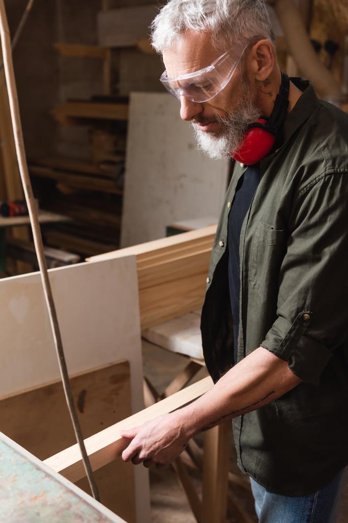 carpinteiro maduro em óculos segurando prancha de madeira no estúdio de madeira - Foto, Imagem