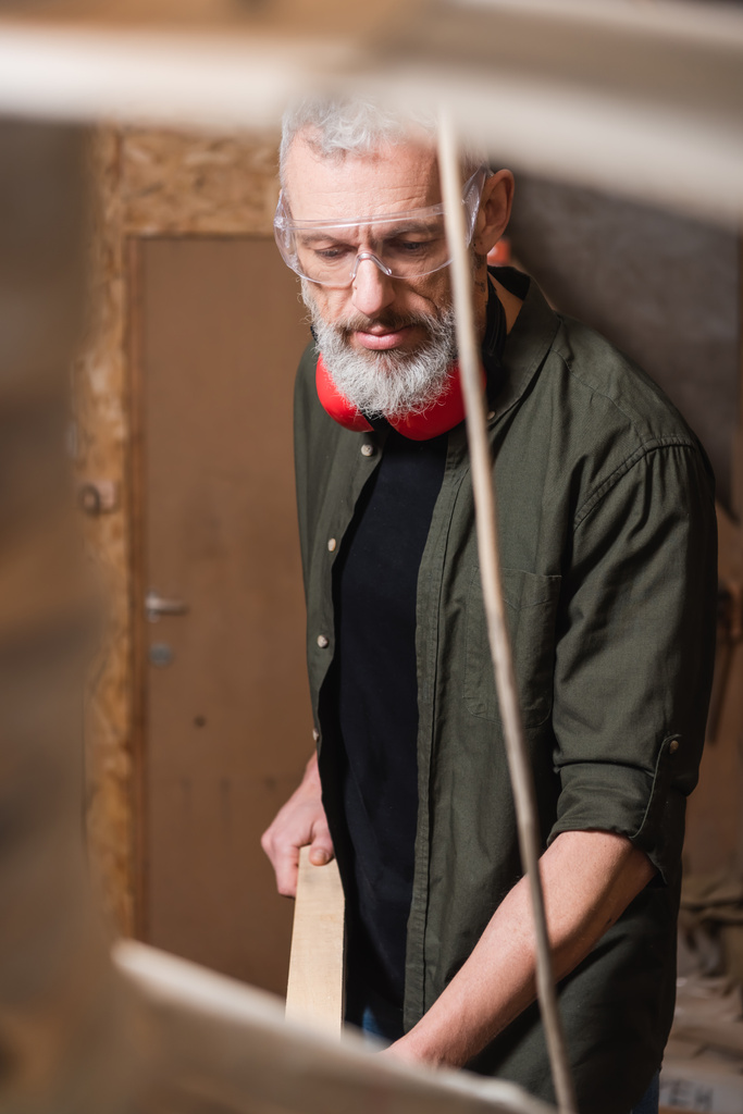 Gözlüklü sakallı mobilya tasarımcısı bulanık ön planda ahşap iş stüdyosunda çalışıyor. - Fotoğraf, Görsel