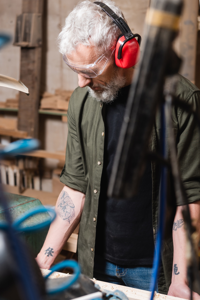 татуйований дизайнер меблів в окулярах і захисних вухах, що працюють в столярній студії
 - Фото, зображення