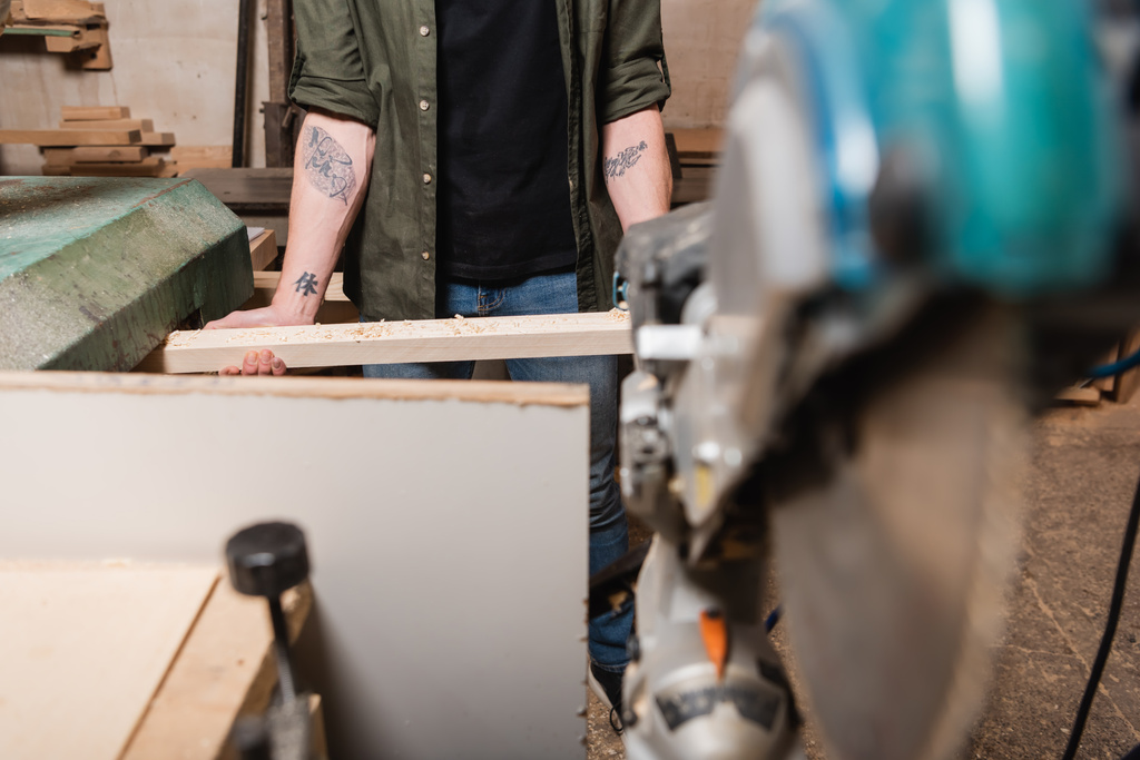 частковий вид татуйованого працівника деревини, що тримає дошку під час роботи на розмитому передньому плані
 - Фото, зображення