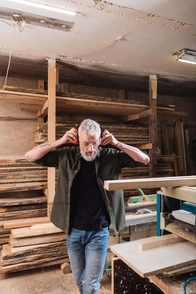 bearded carpenter adjusting protective earmuffs near thickness planer - Photo, Image